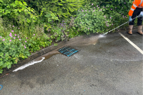 drain in a car park with a field engineer jet washing into the drain. greenery on the bank next to the drain